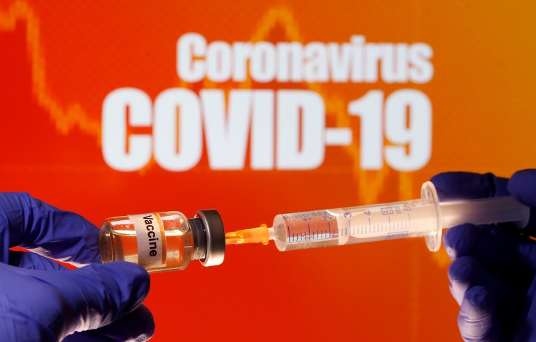 A small bottle labeled with a "Vaccine" sticker is held near a medical syringe in front of displayed "Coronavirus COVID-19" words in this illustration