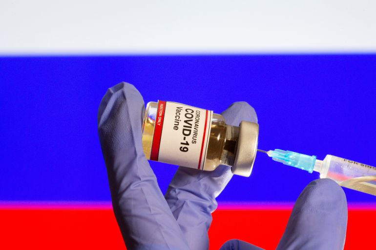A small bottle labeled with a "Coronavirus COVID-19 Vaccine" sticker and a medical syringe in front of displayed Russia flag in this illustration